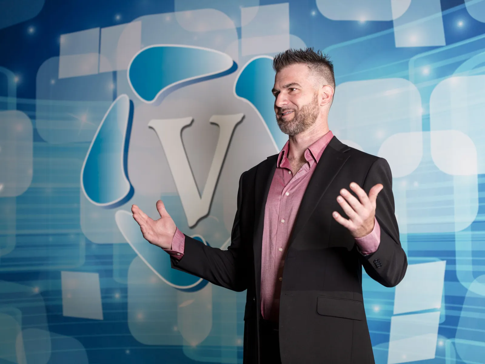 Man in black blazer and pink shirt gesturing with hands in front of blue digital background with a large V logo