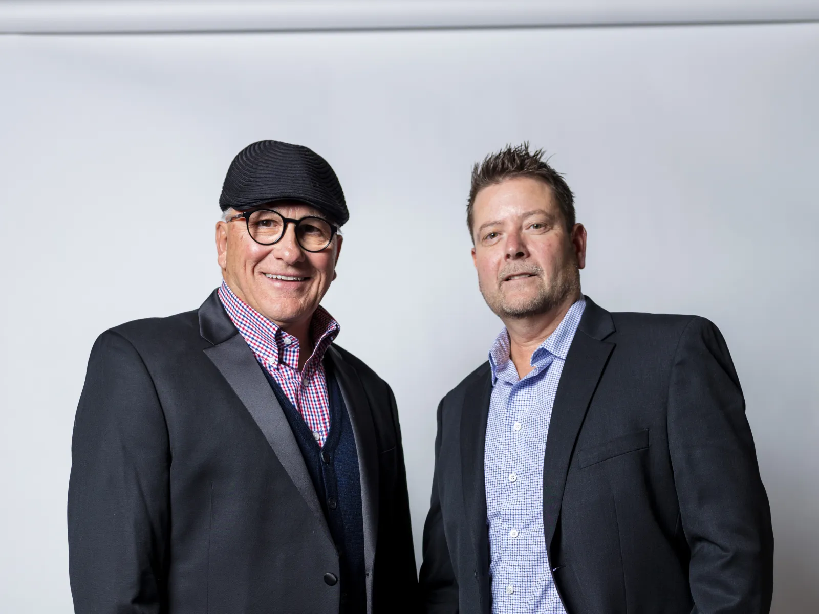 Two men in business attire posing against a white background, one wearing a cap and glasses, the other holding eyeglasses.