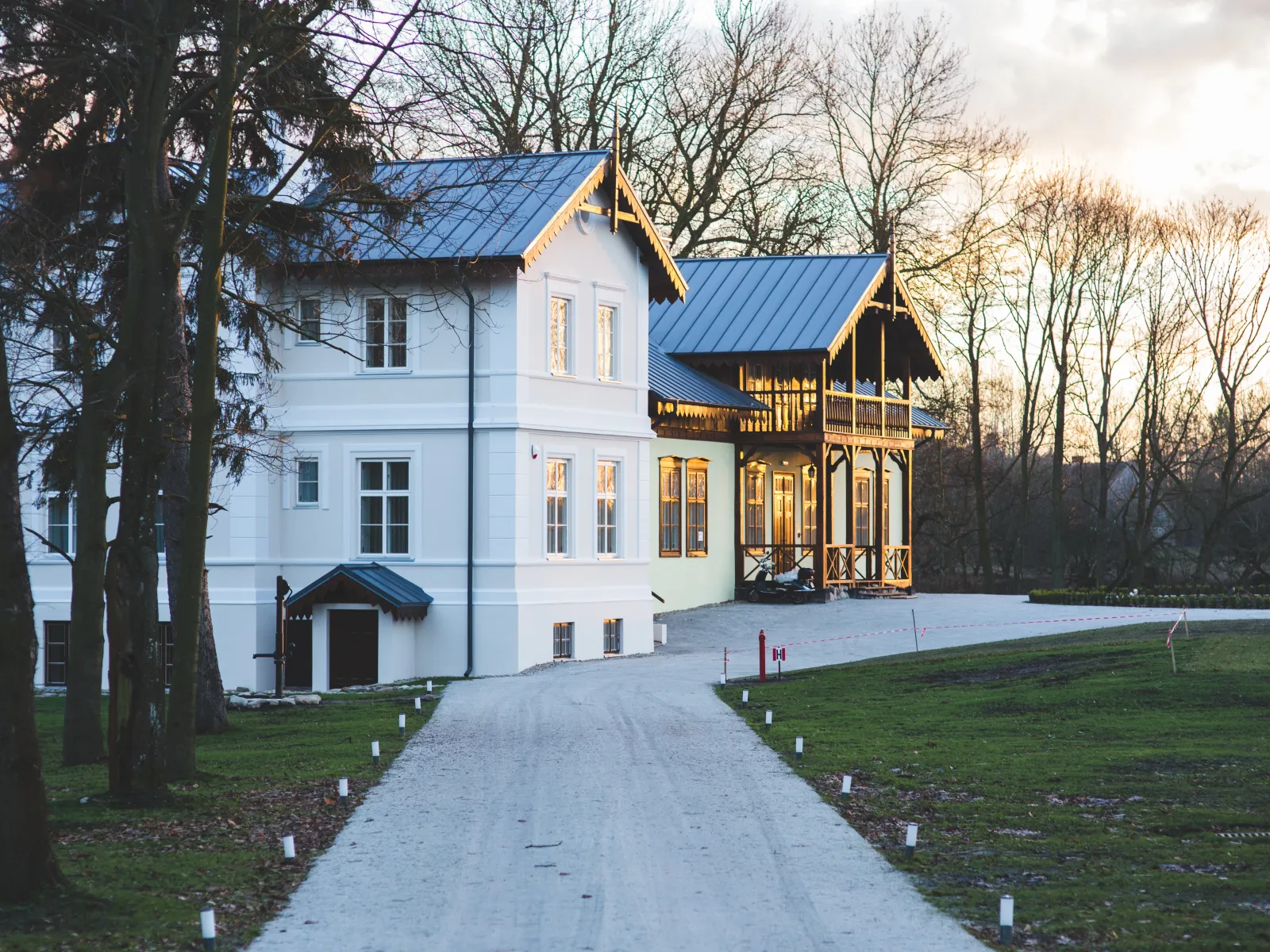 White and beige house with metal roofs along a paved driveway at sunset surrounded by bare trees and grass.