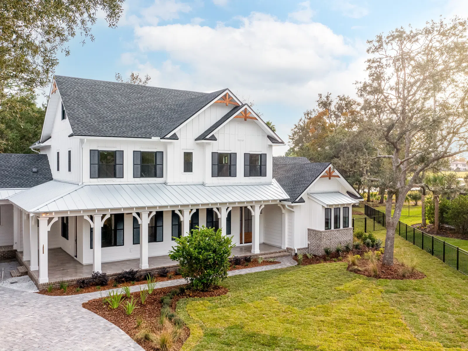 Modern two-story white farmhouse with a wraparound porch, manicured lawn, and surrounding trees in bright daylight