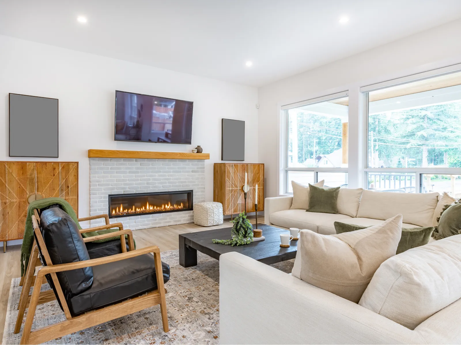 Bright modern living room with large windows, beige sofas, black leather chair, fireplace, and wooden cabinets.