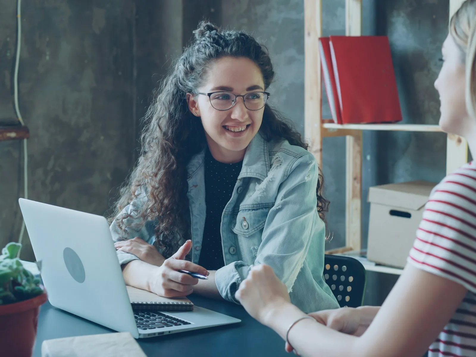 Two women engaged in a friendly conversation at a table with a laptop and notebooks in a casual workspace.