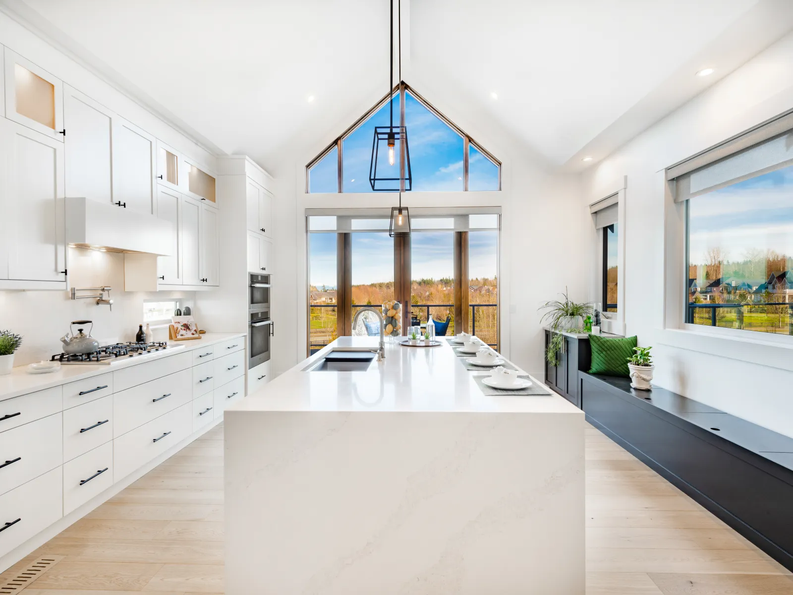 Bright modern kitchen with large white island, vaulted windows, black fixtures, and scenic outdoor views.