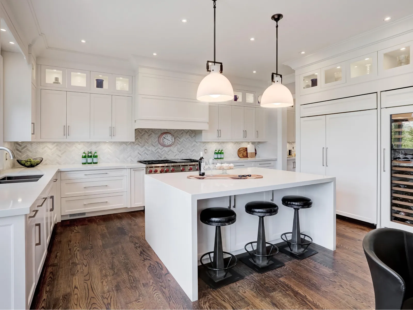 Modern white kitchen with large island, black bar stools, pendant lights, hardwood floors, and built-in wine cooler.