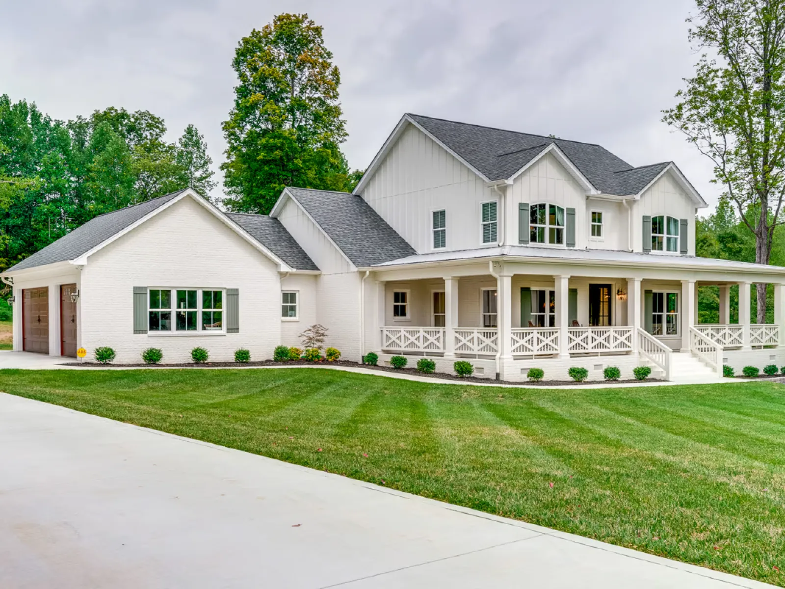 Charming modern farmhouse with large porch, manicured lawn, and beautiful landscaping in a serene setting.