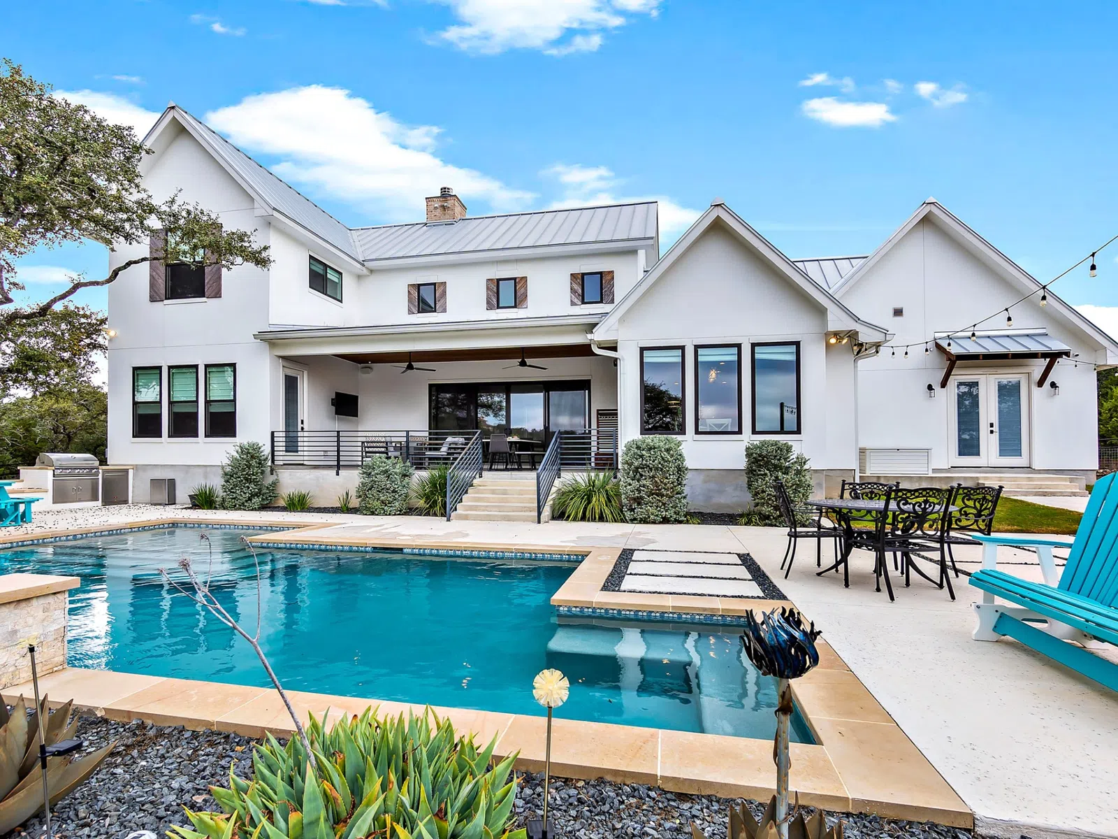 Modern white house with a backyard swimming pool, blue lounge chairs, dining area, and clear sky.