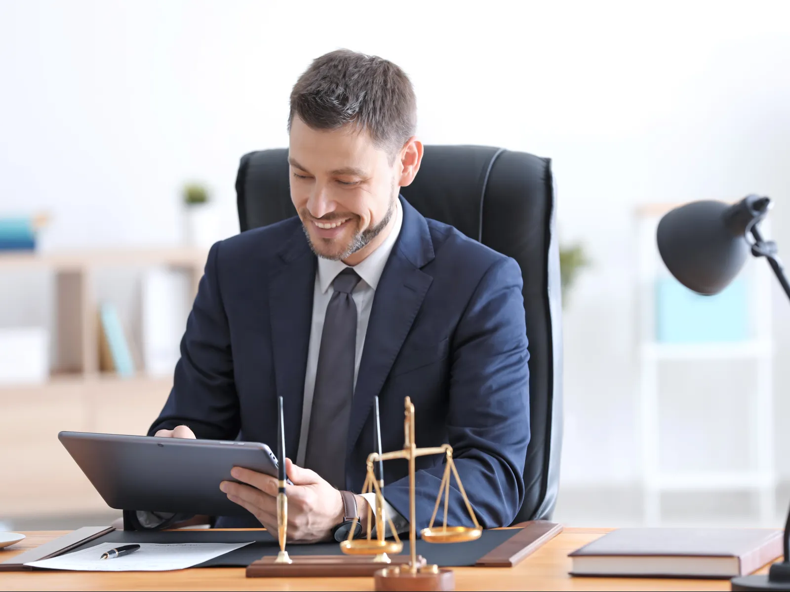Smiling businessman in suit working on tablet at desk with legal scales and documents in modern office.