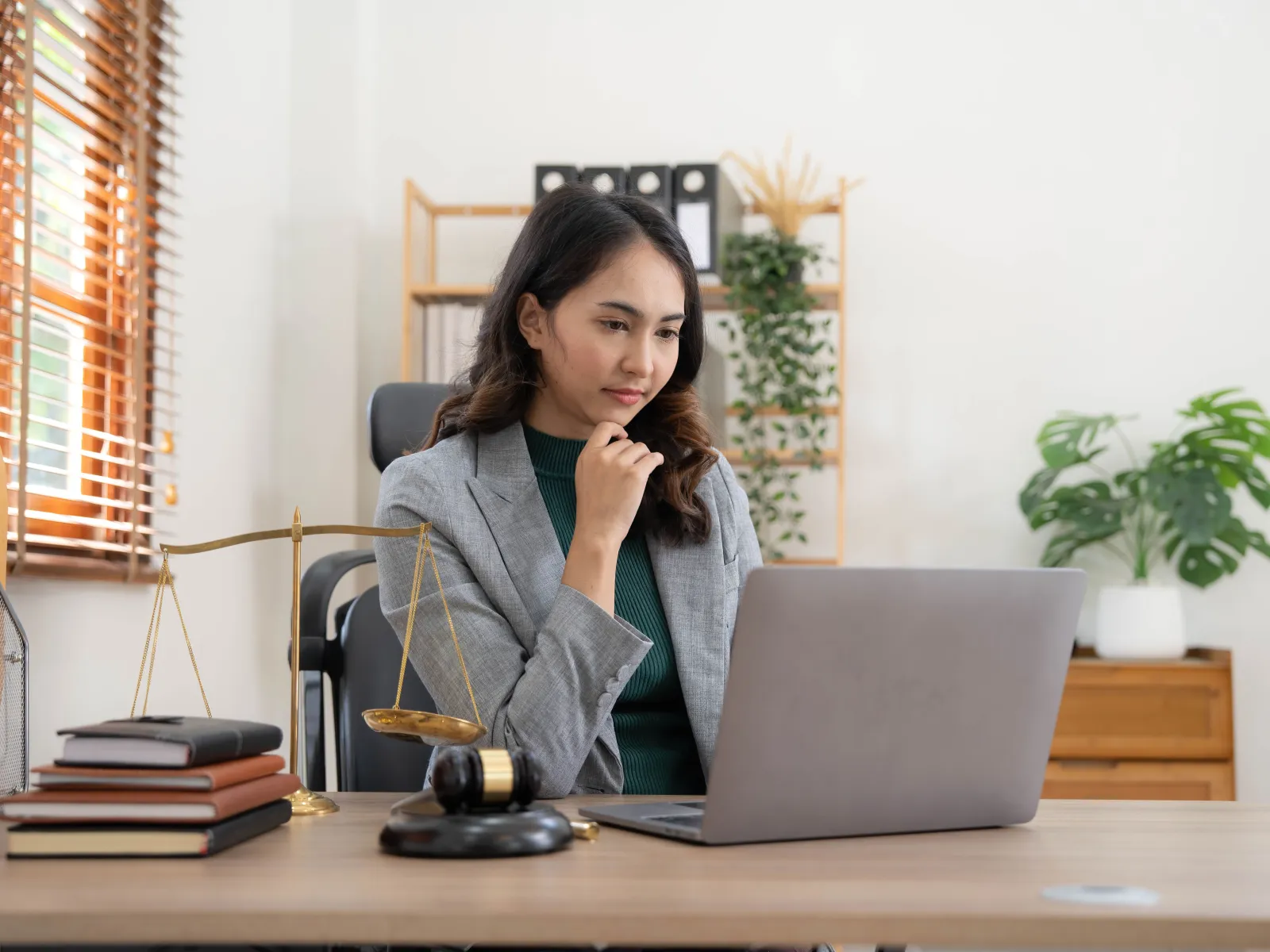 Woman in a gray blazer working on a laptop at a desk with legal books, scales of justice, and a gavel.