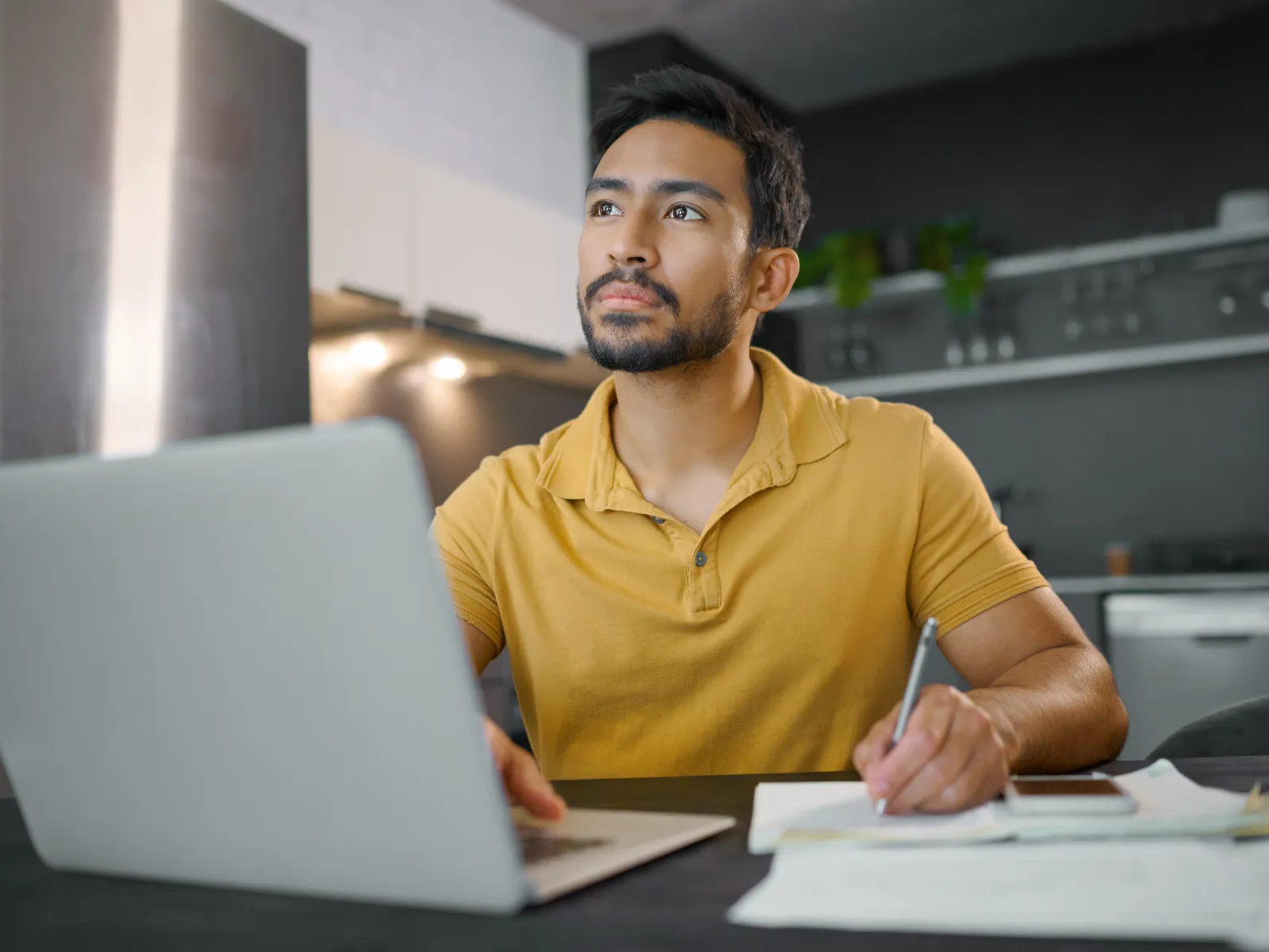 Man in yellow shirt working on laptop and taking notes at home kitchen workspace.