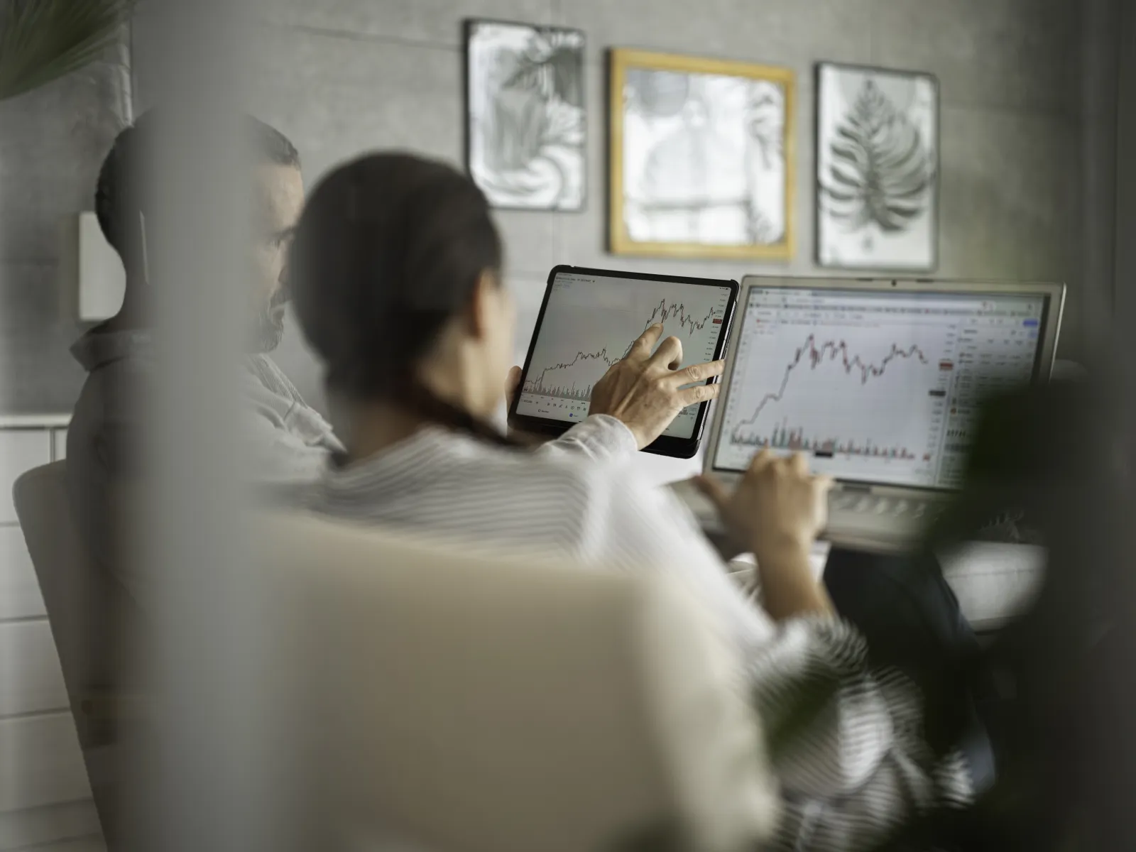 Two professionals analyzing financial charts on laptop and tablet in a modern office with framed art.
