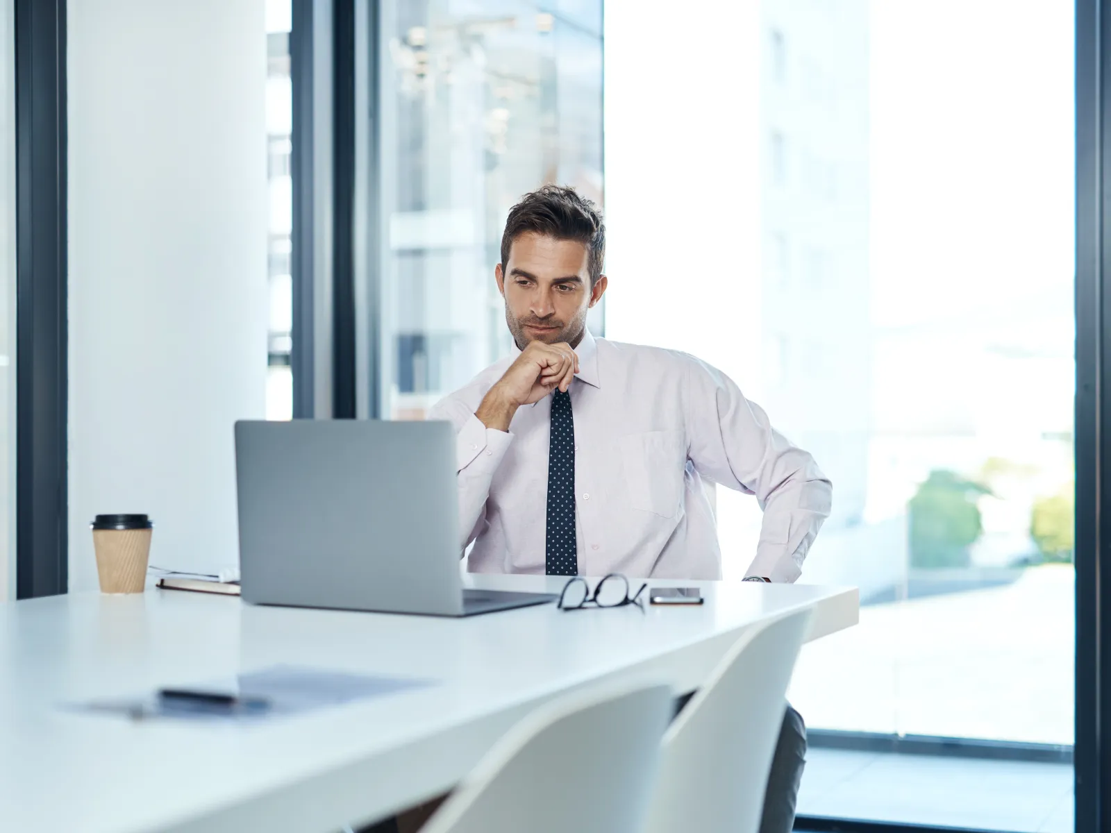 Businessman in white shirt working on laptop in modern office with large windows and natural light