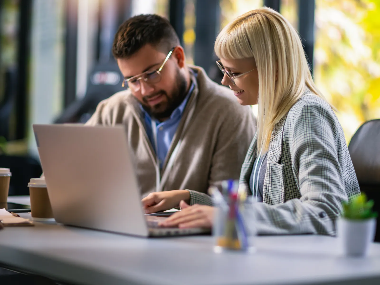 Two colleagues working together on a laptop in a bright modern office with coffee cups and office supplies.
