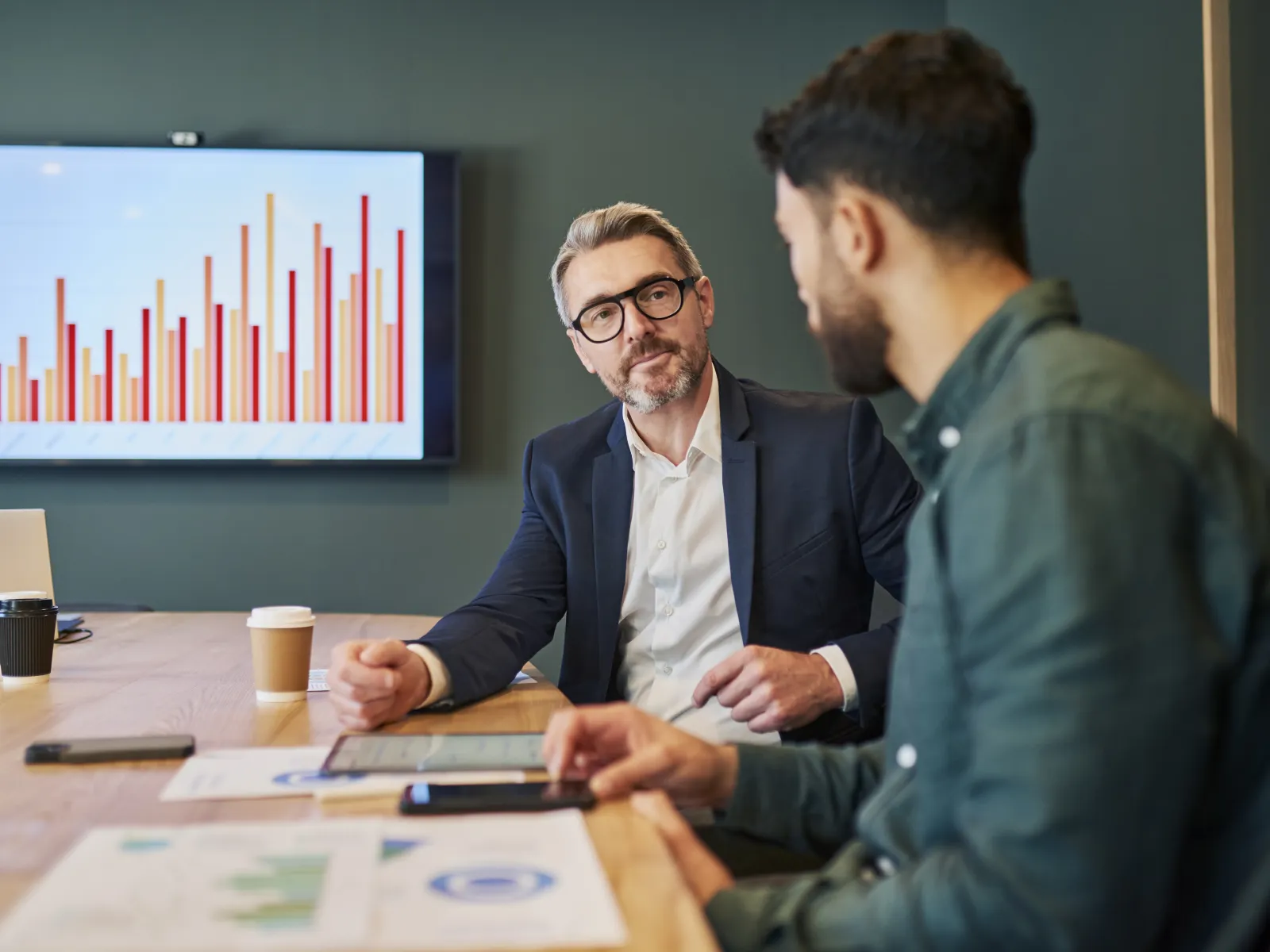 Two businessmen discussing documents and digital data in office meeting with bar chart on screen in background