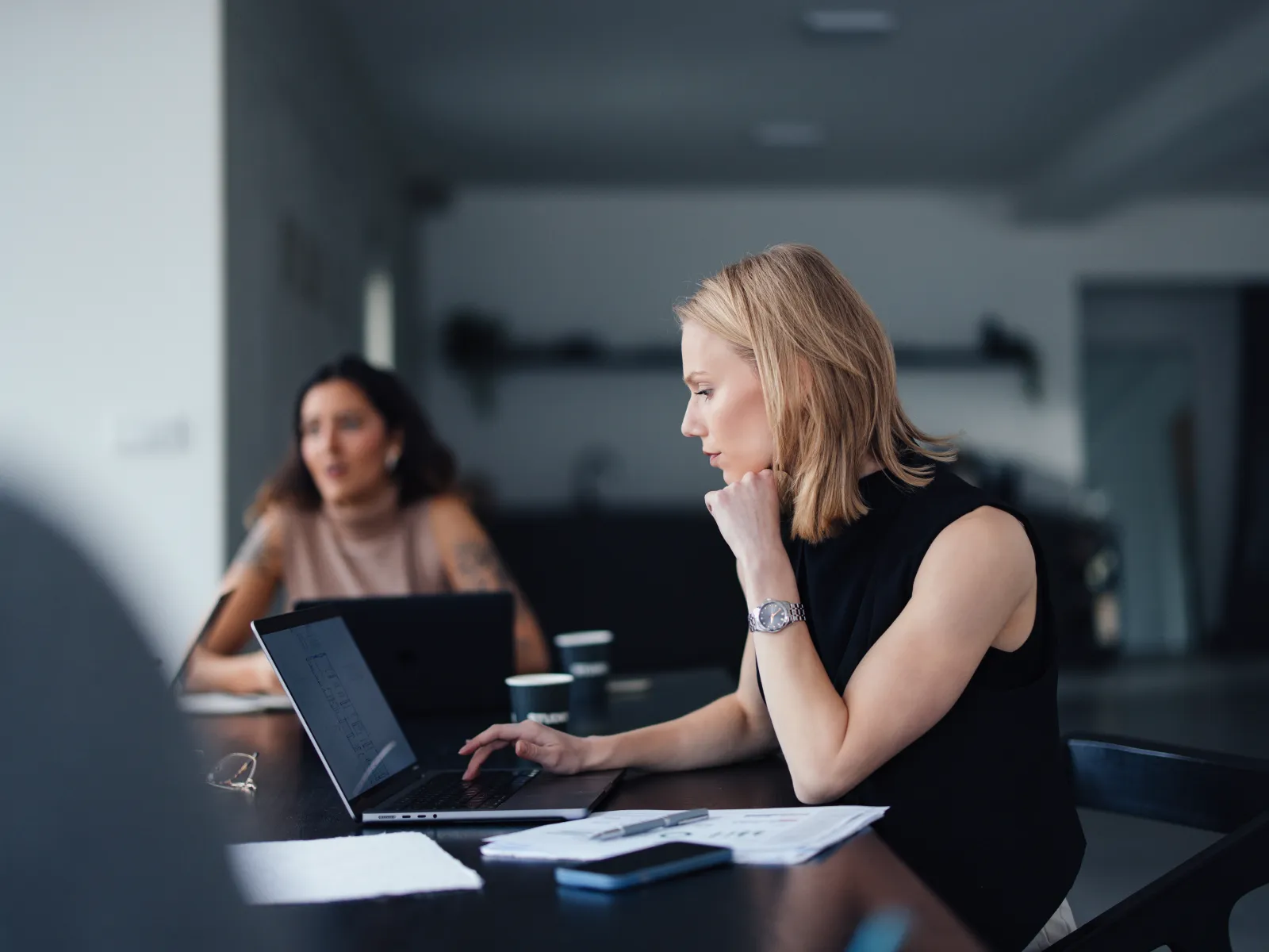 Focused woman working on laptop at meeting table with documents and colleagues in modern office environment