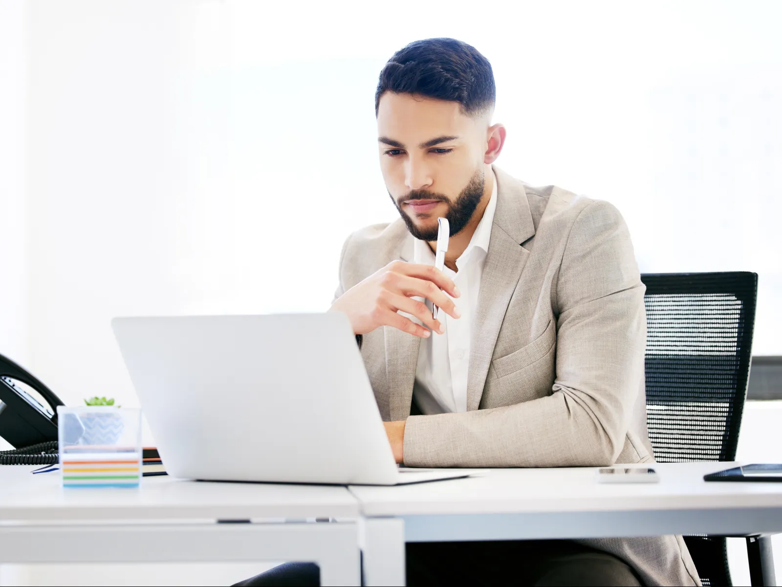 Young businessman thoughtfully working on laptop at office desk with phone and plant nearby