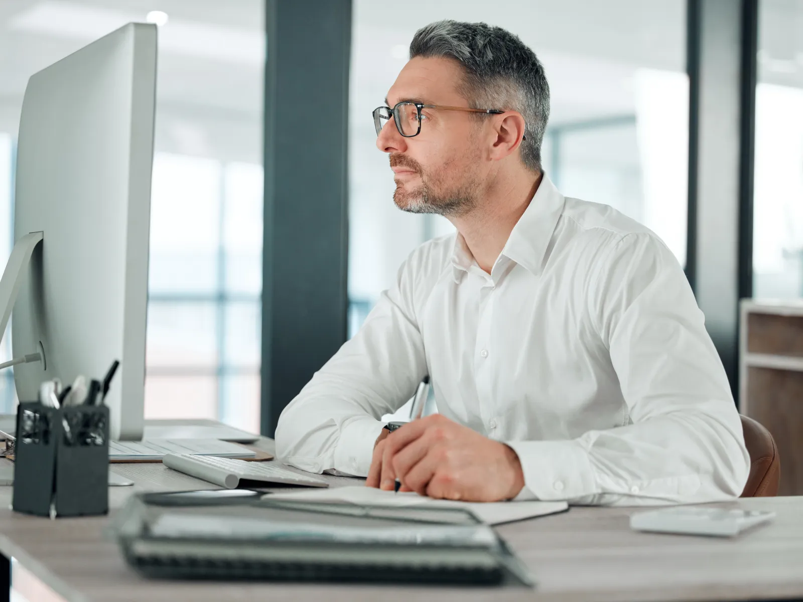 Man in white shirt and glasses working focused at a desktop computer in a bright modern office.