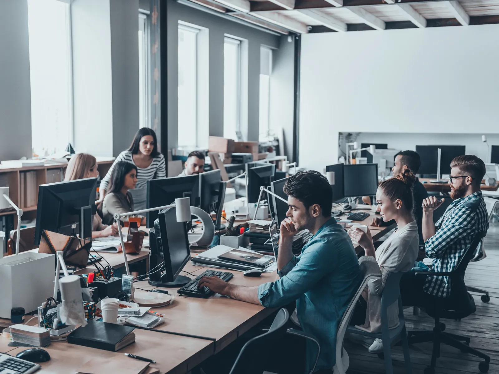 Diverse team working on computers in a modern open office with large windows and wooden ceiling beams
