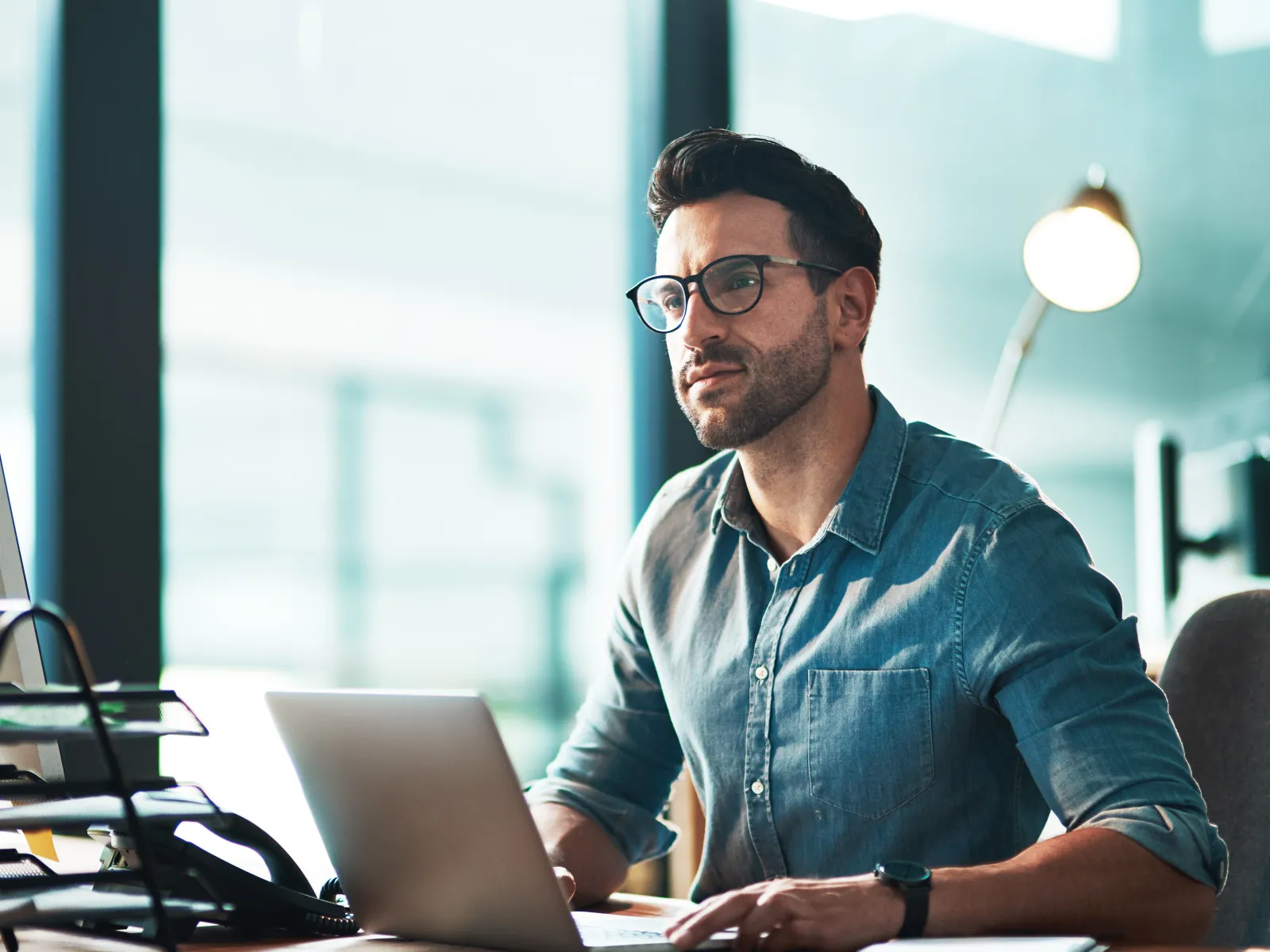 Man wearing glasses working on a laptop at a desk in a modern office with natural light.