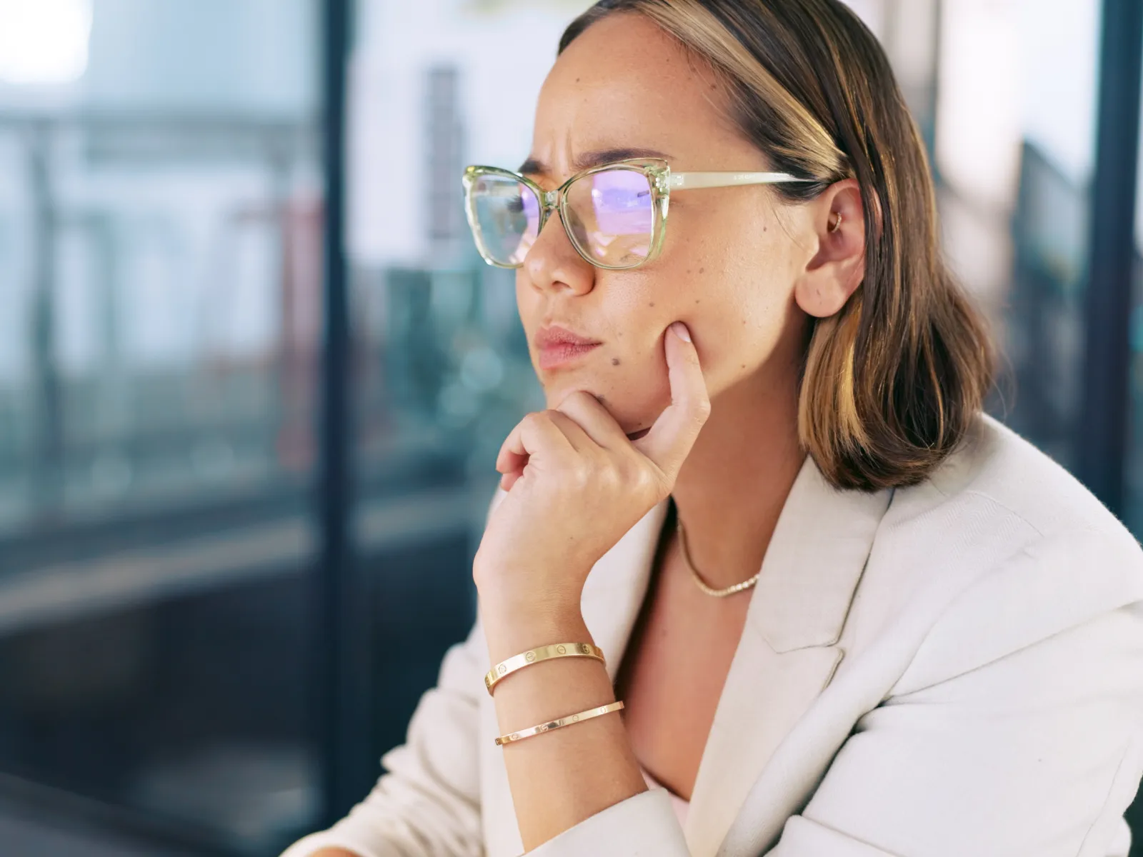 Young woman wearing glasses, focused and thinking at her desk in a modern office setting.