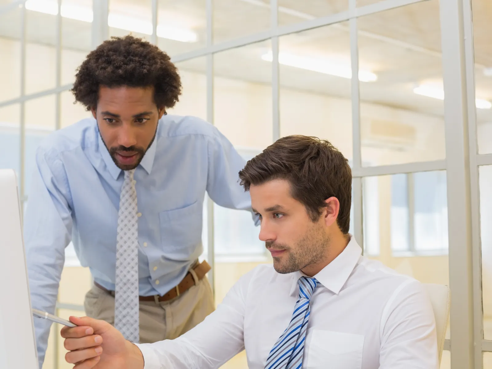 Two male colleagues in business attire collaborating and discussing work on a desktop computer in an office.