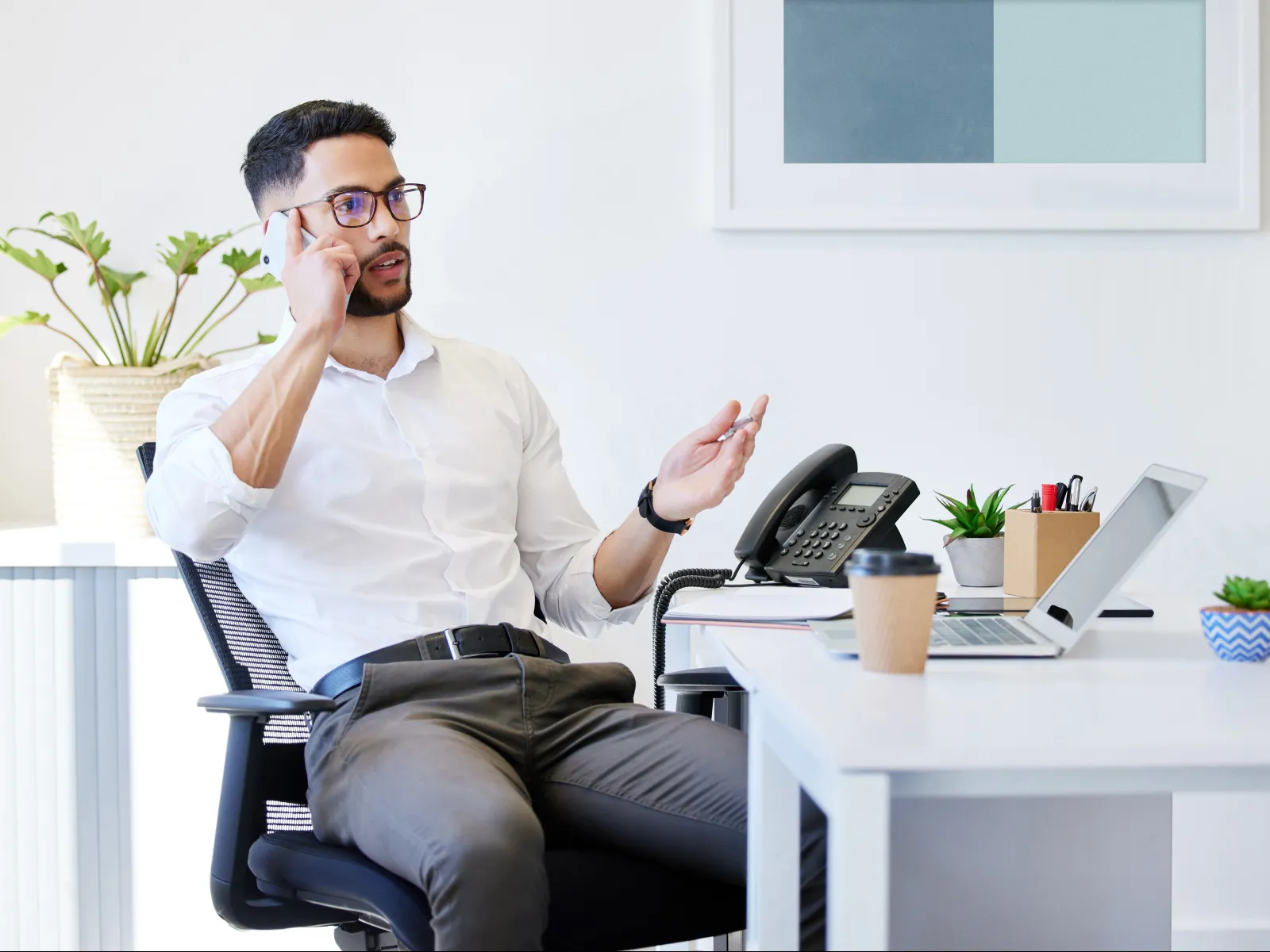 Man in white shirt and glasses talking on phone in a modern bright office with laptop and plants.