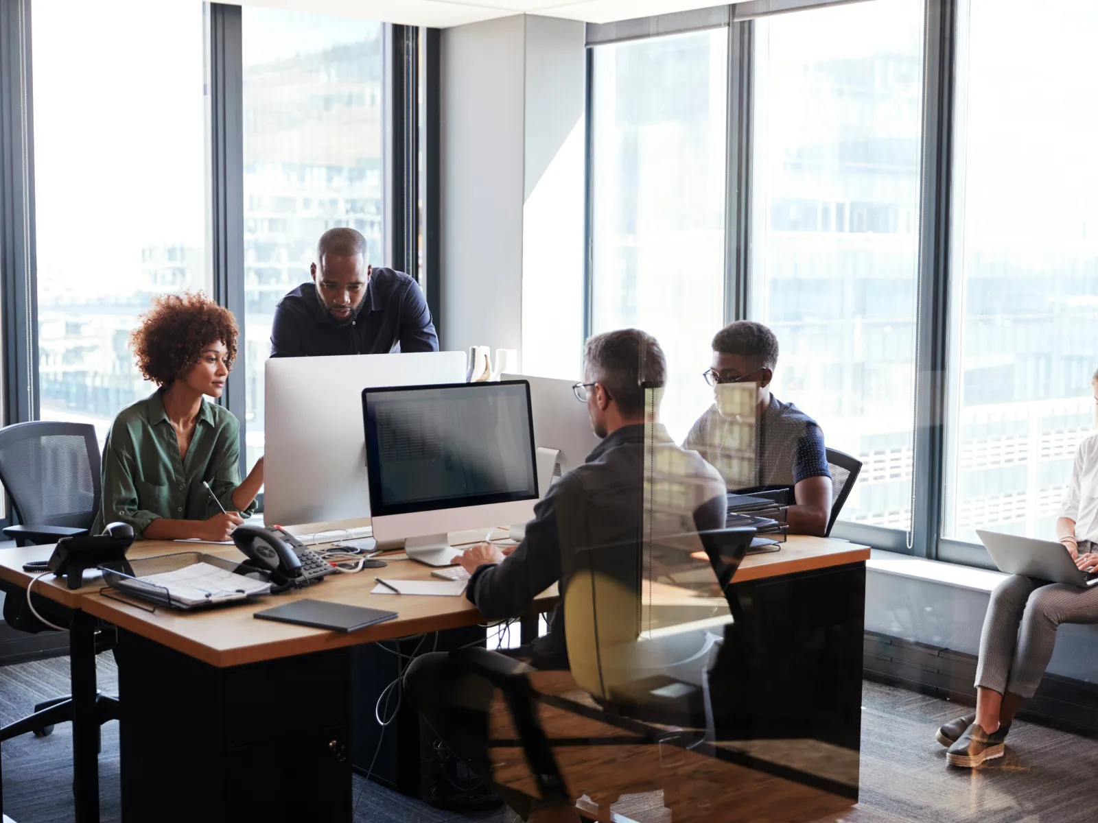 Diverse office team working together at desks with computers in a modern bright workspace with large windows.