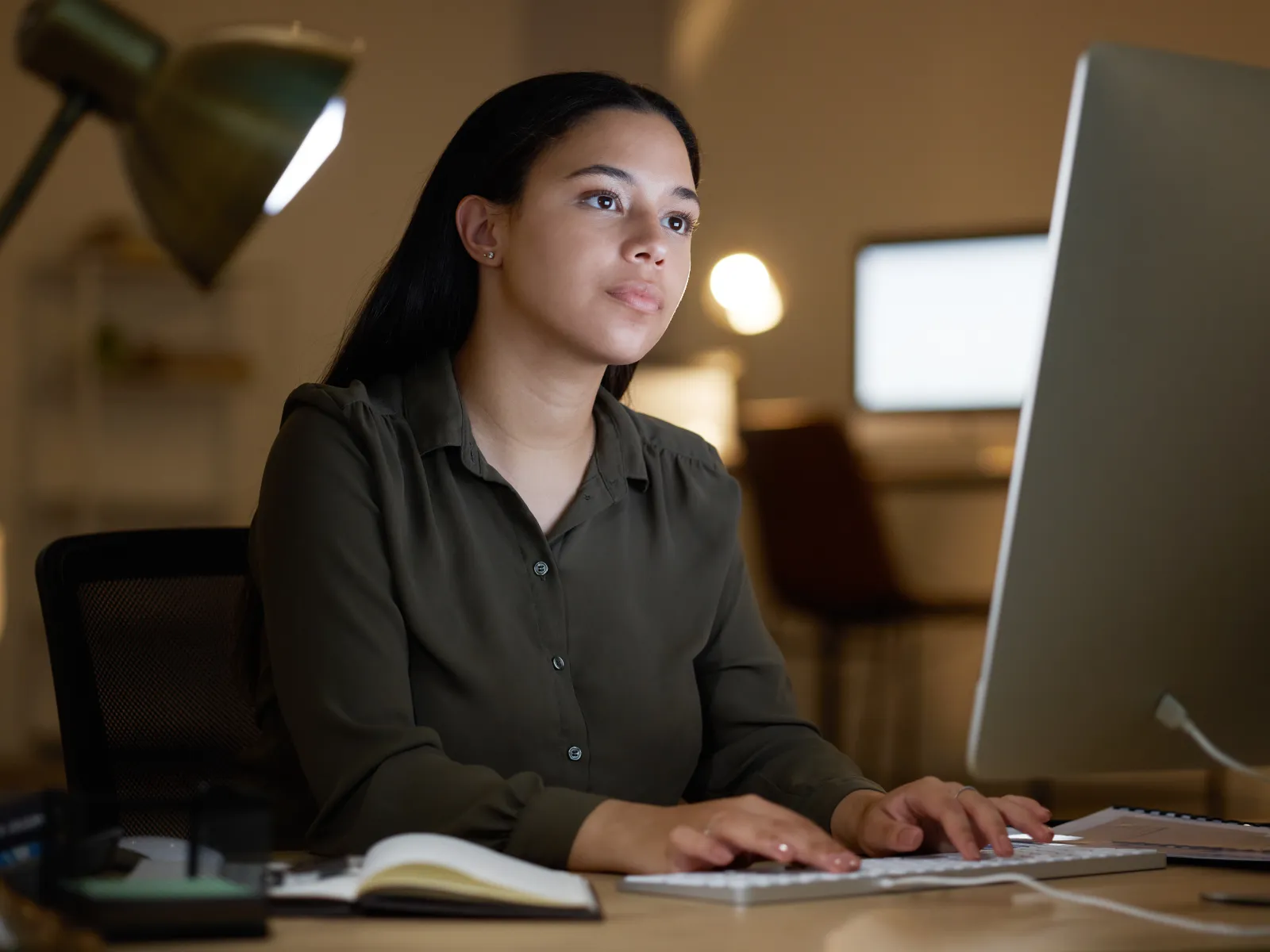Young woman working late on a computer at a desk with lamp and notebook in a cozy office setting