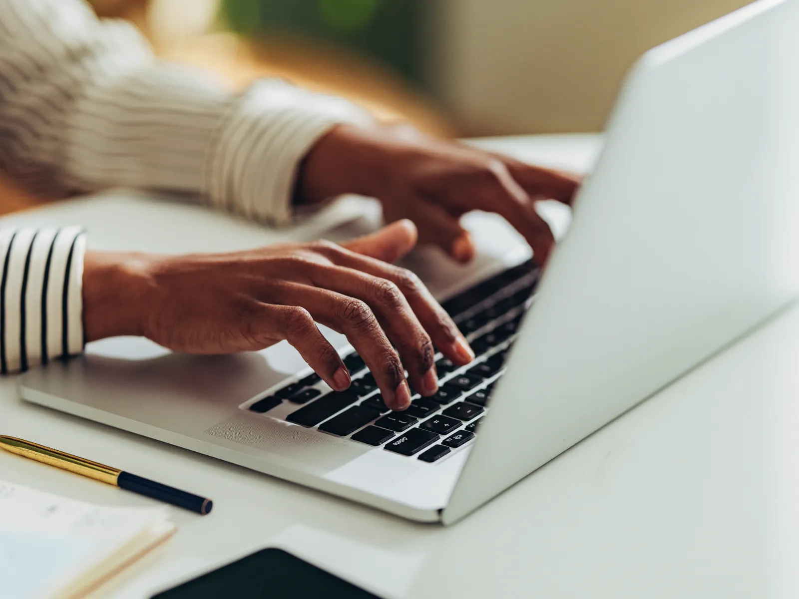 Close-up of hands typing on a laptop keyboard on a white desk with a smartphone and pen nearby
