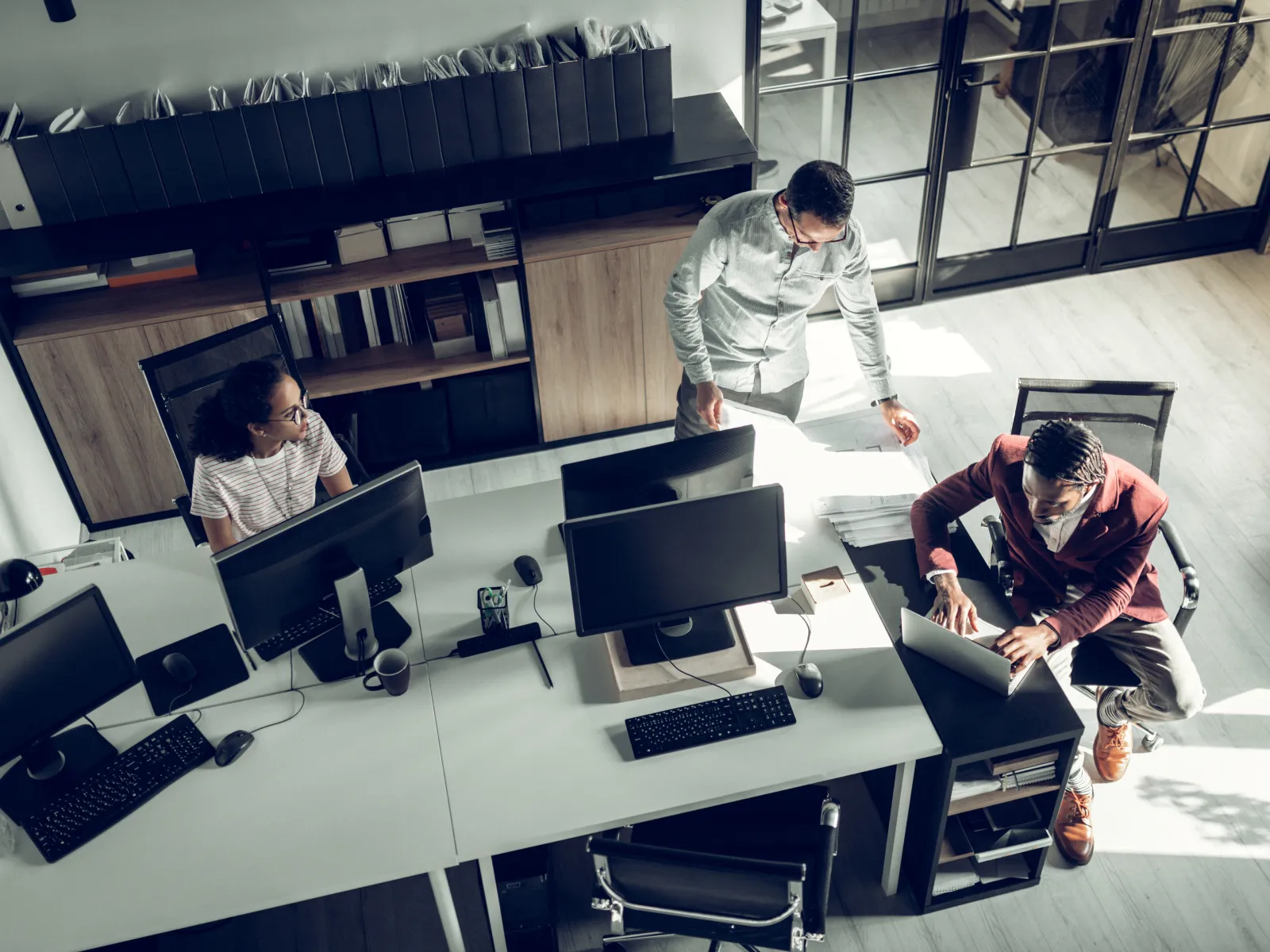 Diverse office team working together at desks with computers in a modern bright workspace with large windows.