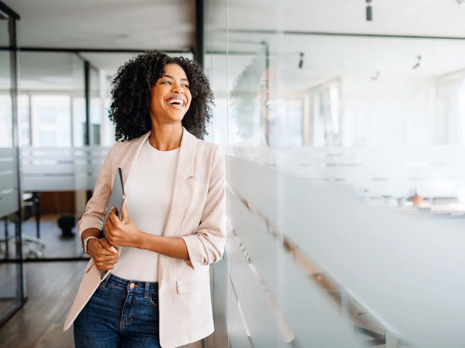Smiling businesswoman in casual blazer holding tablet in modern office with glass walls and natural light