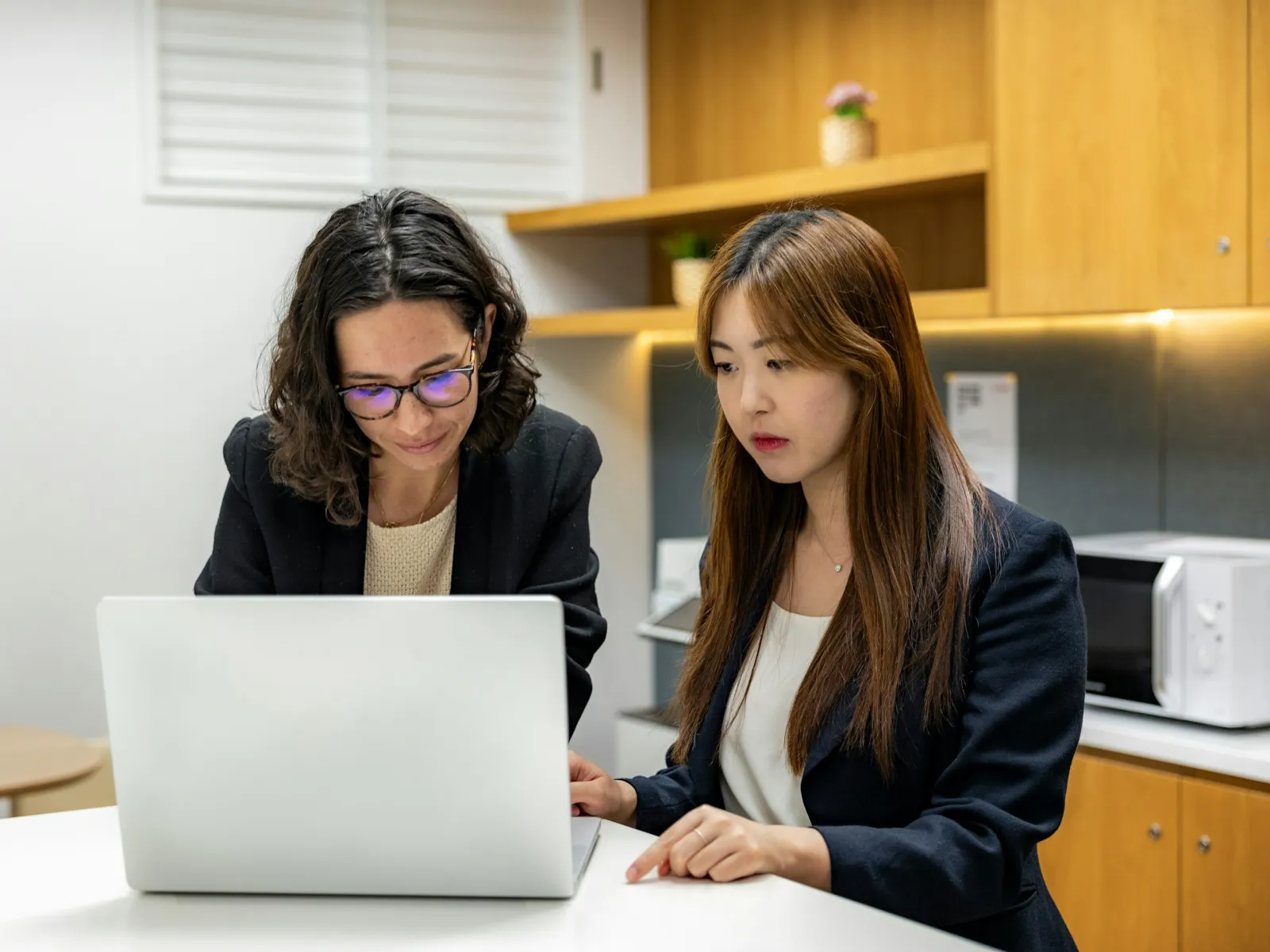 Two businesswomen collaborating and working together on a laptop at a modern office kitchen space.