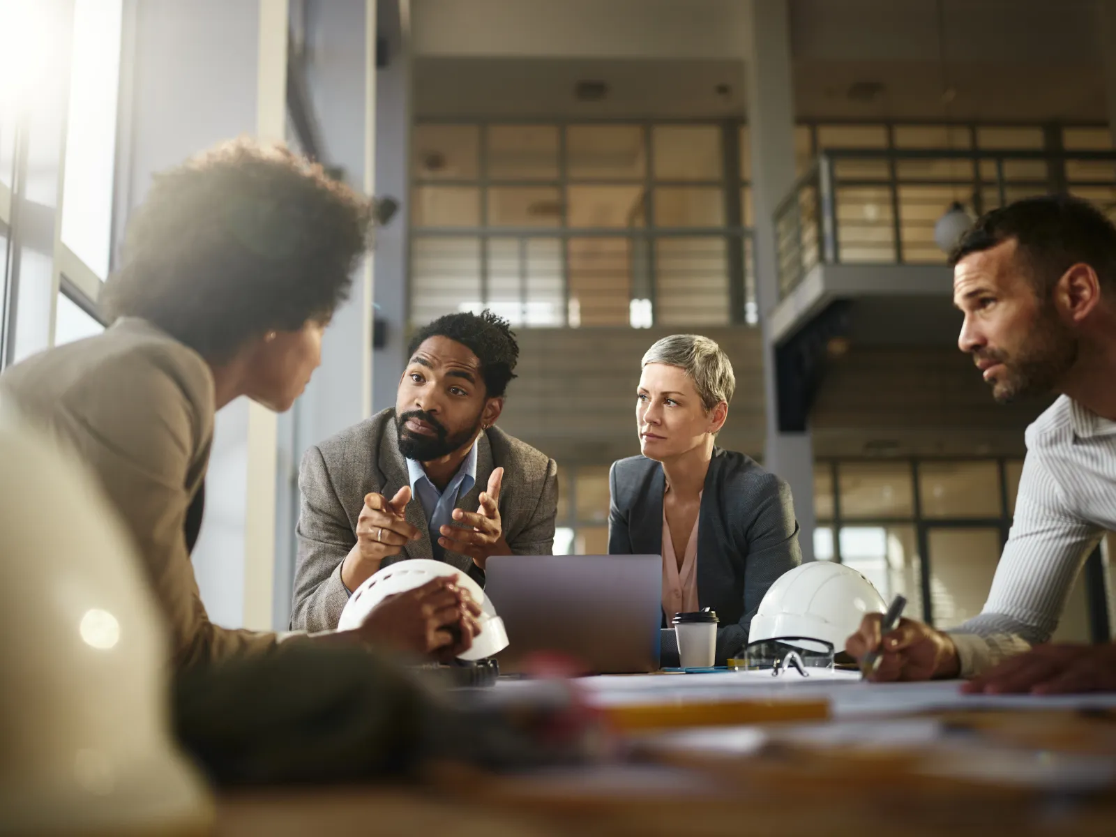 Business professionals wearing blazers discuss a project around a table with safety helmets in a modern office.
