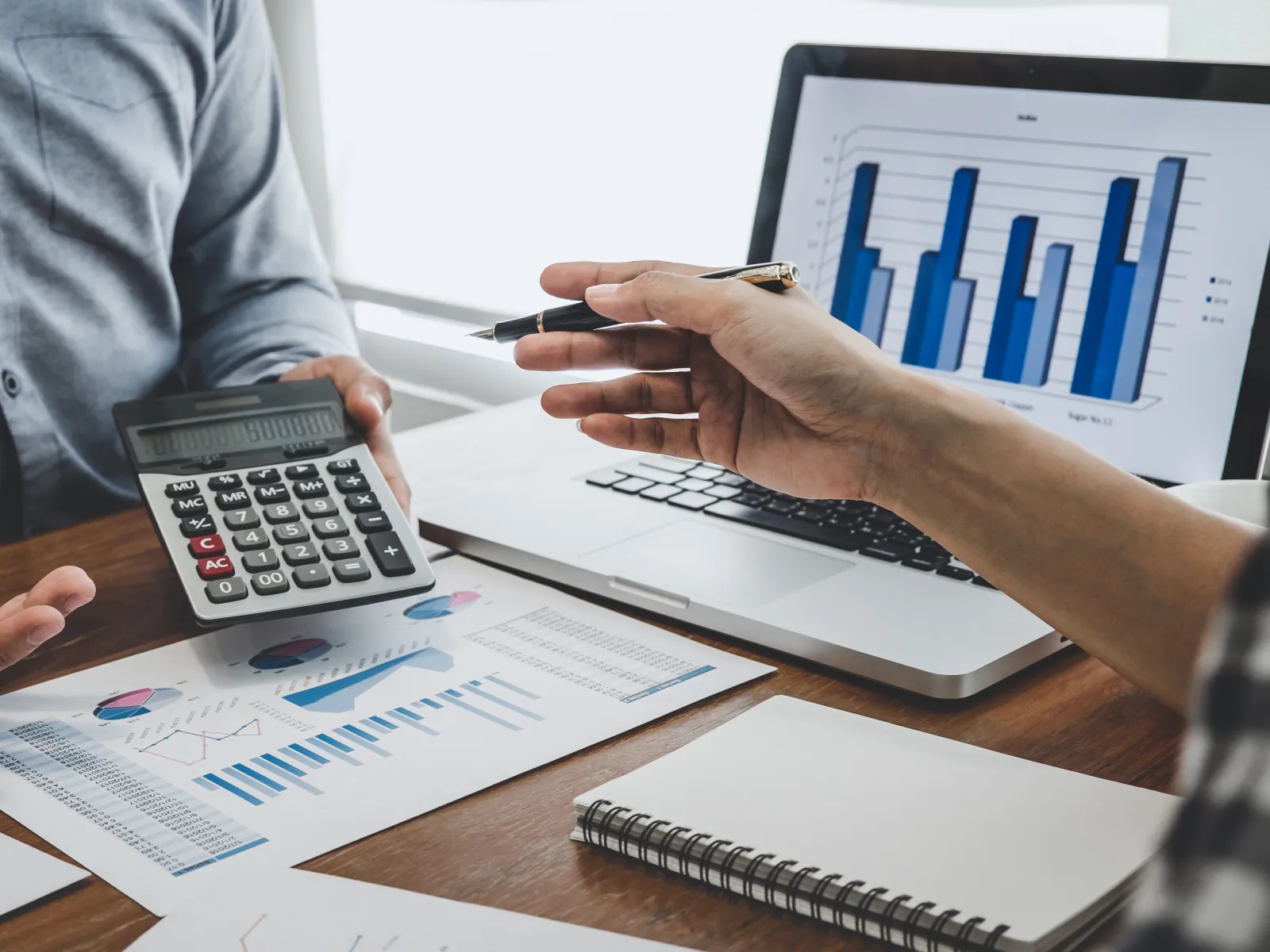 Two people discussing financial charts and data using a calculator and laptop with bar graphs displayed