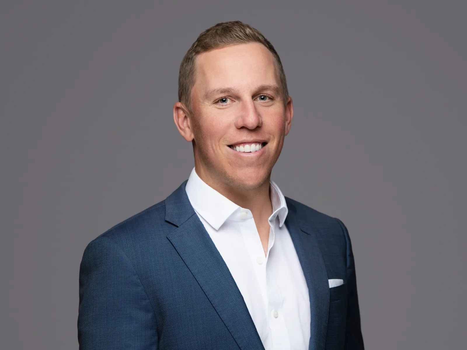Professional portrait of a smiling man in a navy blue suit and white dress shirt against a gray background