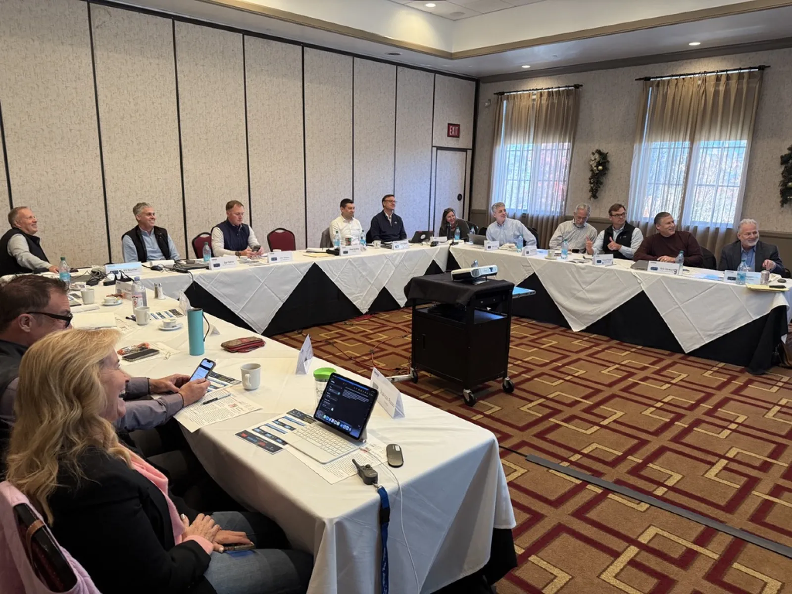 Business professionals seated around a U-shaped conference table in a meeting room with a projector and laptops.