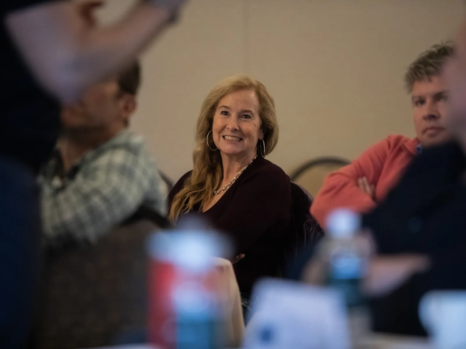 Smiling woman with long hair sitting among a group in a meeting setting with blurred foreground objects.