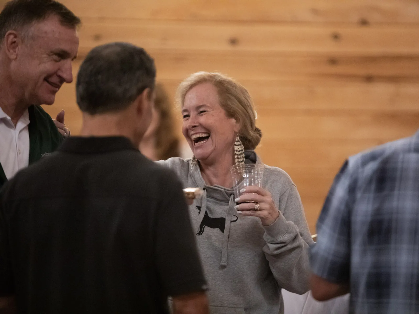 Group of adults socializing indoors, laughing and holding drinks in a casual setting with warm wooden background.