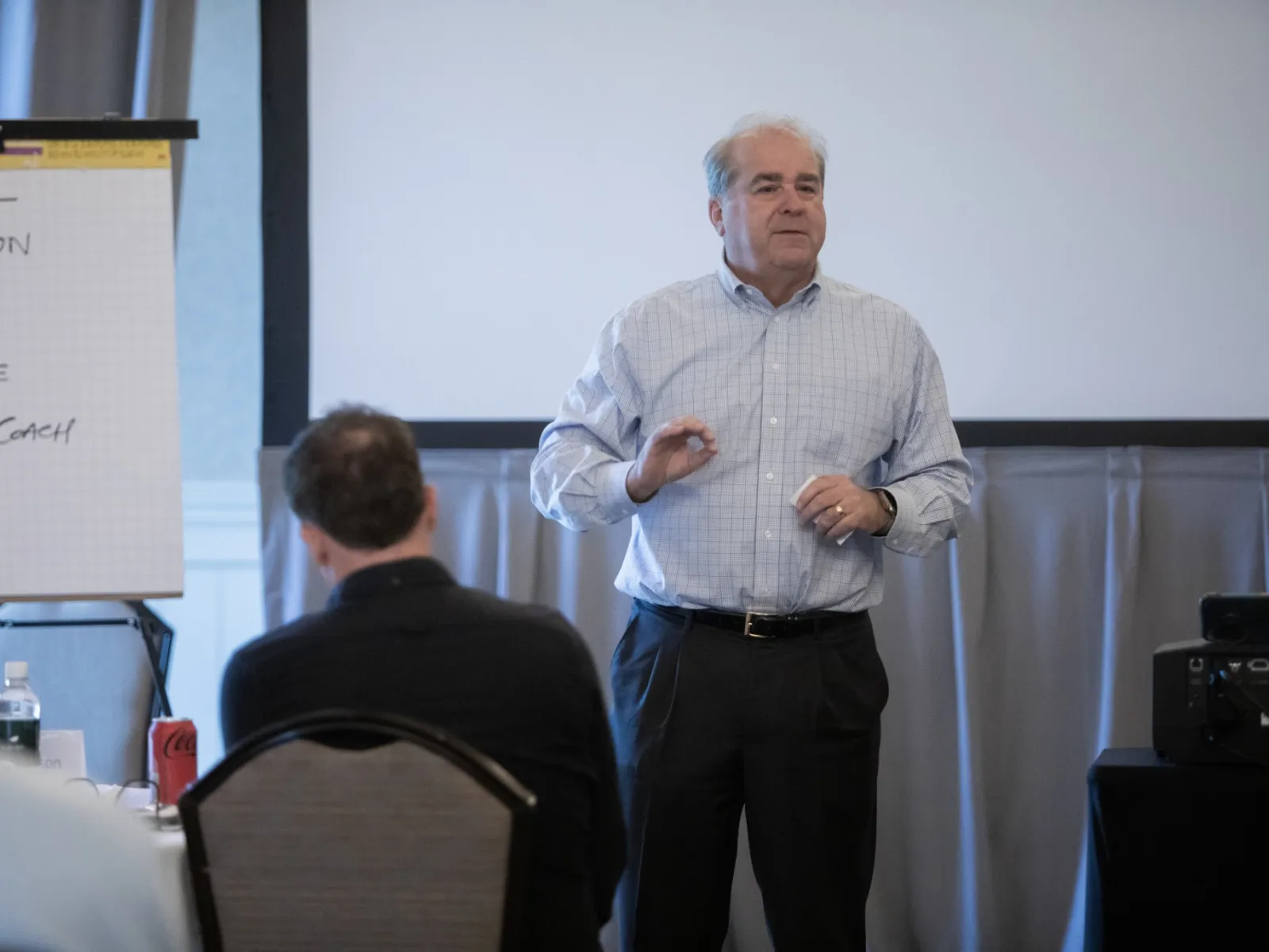 Man in plaid shirt writing on large flip chart with marker during a presentation or meeting indoors.