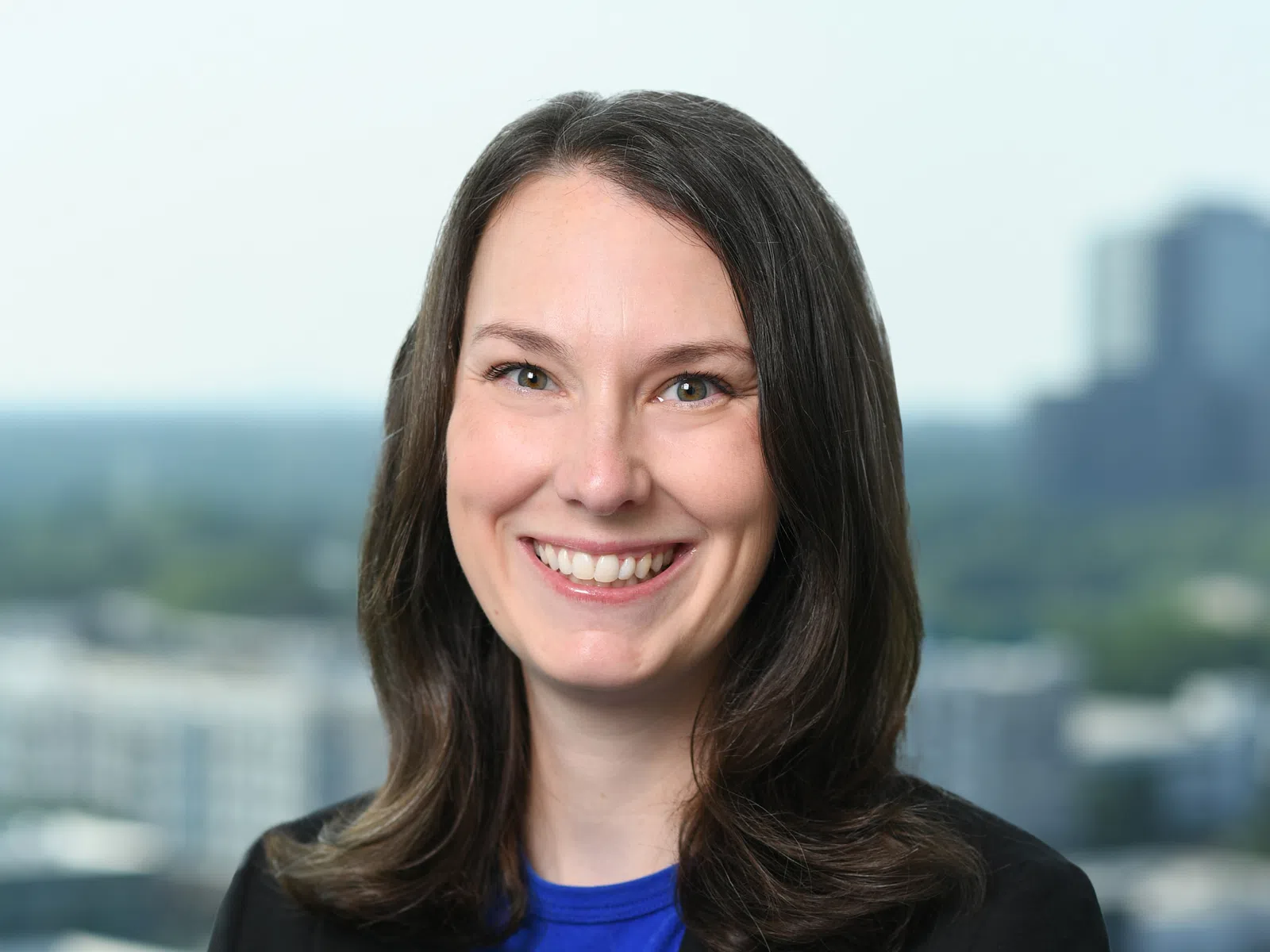 Smiling woman with shoulder-length dark hair wearing a blue top and black blazer against a cityscape background.