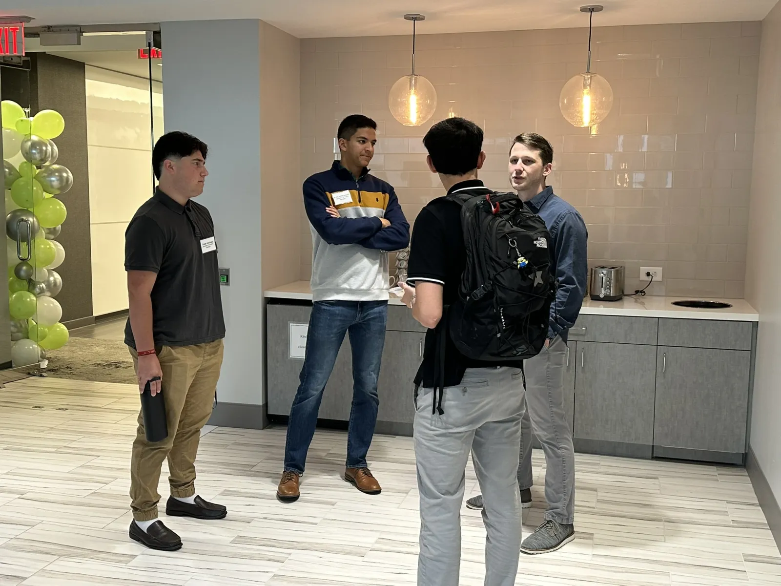 Four young men engaged in conversation in a modern office kitchen area with pendant lights and balloons.