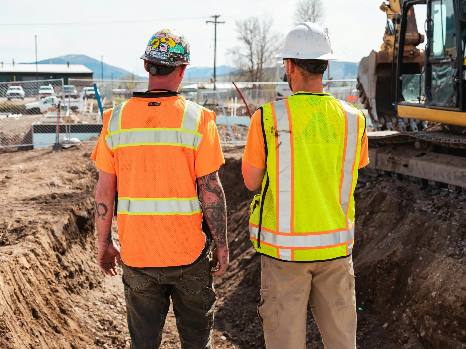Two construction workers in safety vests and helmets standing by an excavation site with heavy machinery.