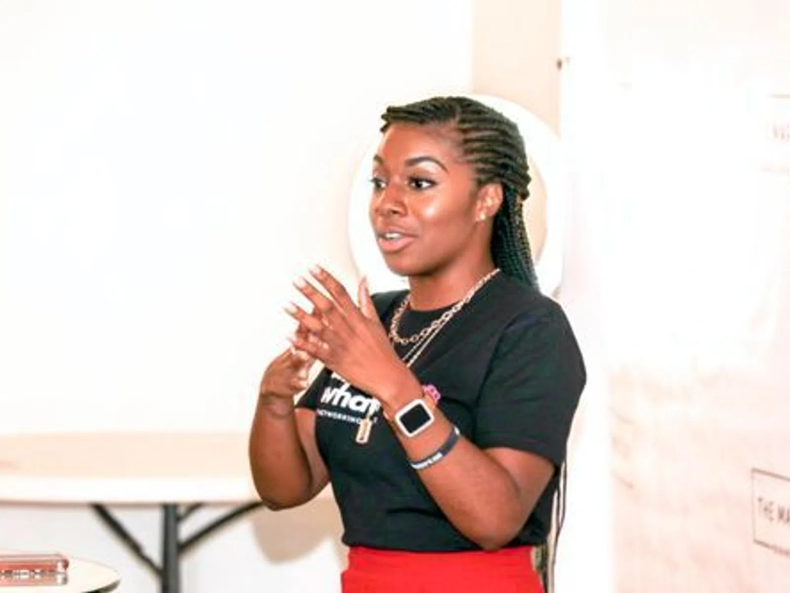 Young woman with braided hair speaking and gesturing with hands indoors wearing a black shirt and red skirt.