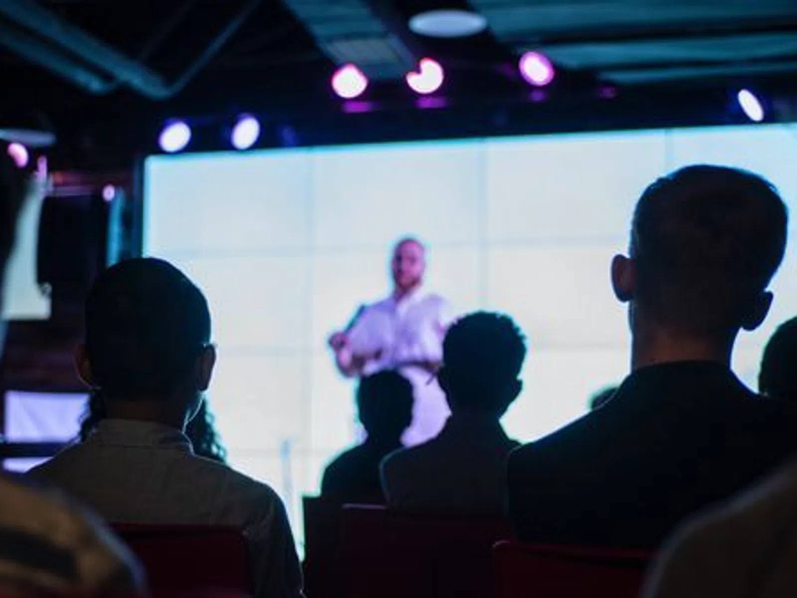 Audience watching a speaker present on stage with a large screen and purple stage lights in a dark room.