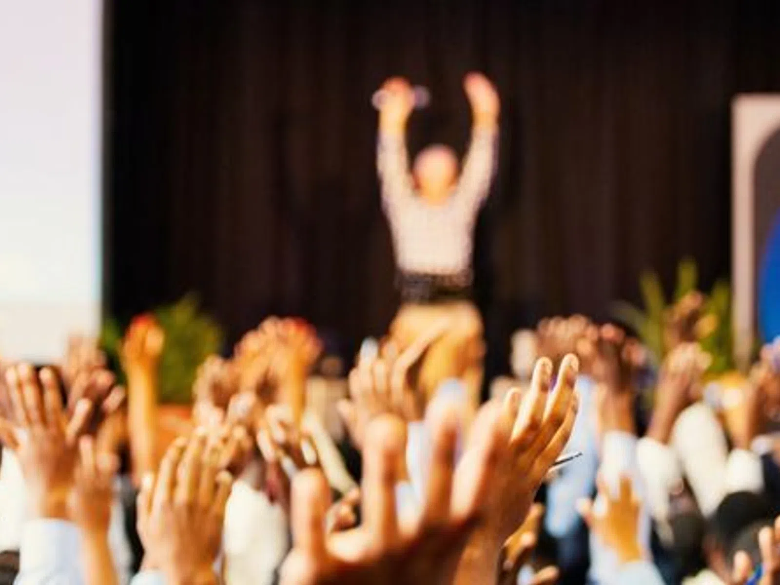Crowd raising hands enthusiastically during a lively event with a speaker in the background on stage.