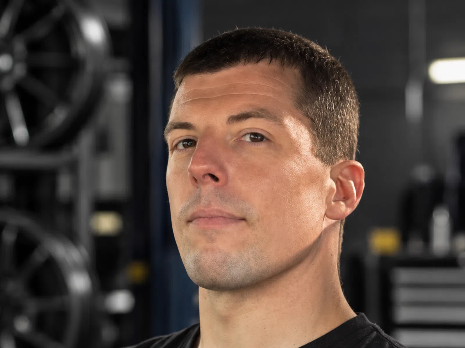 Confident man in black shirt with crossed arms in an industrial setting with machinery in the background.