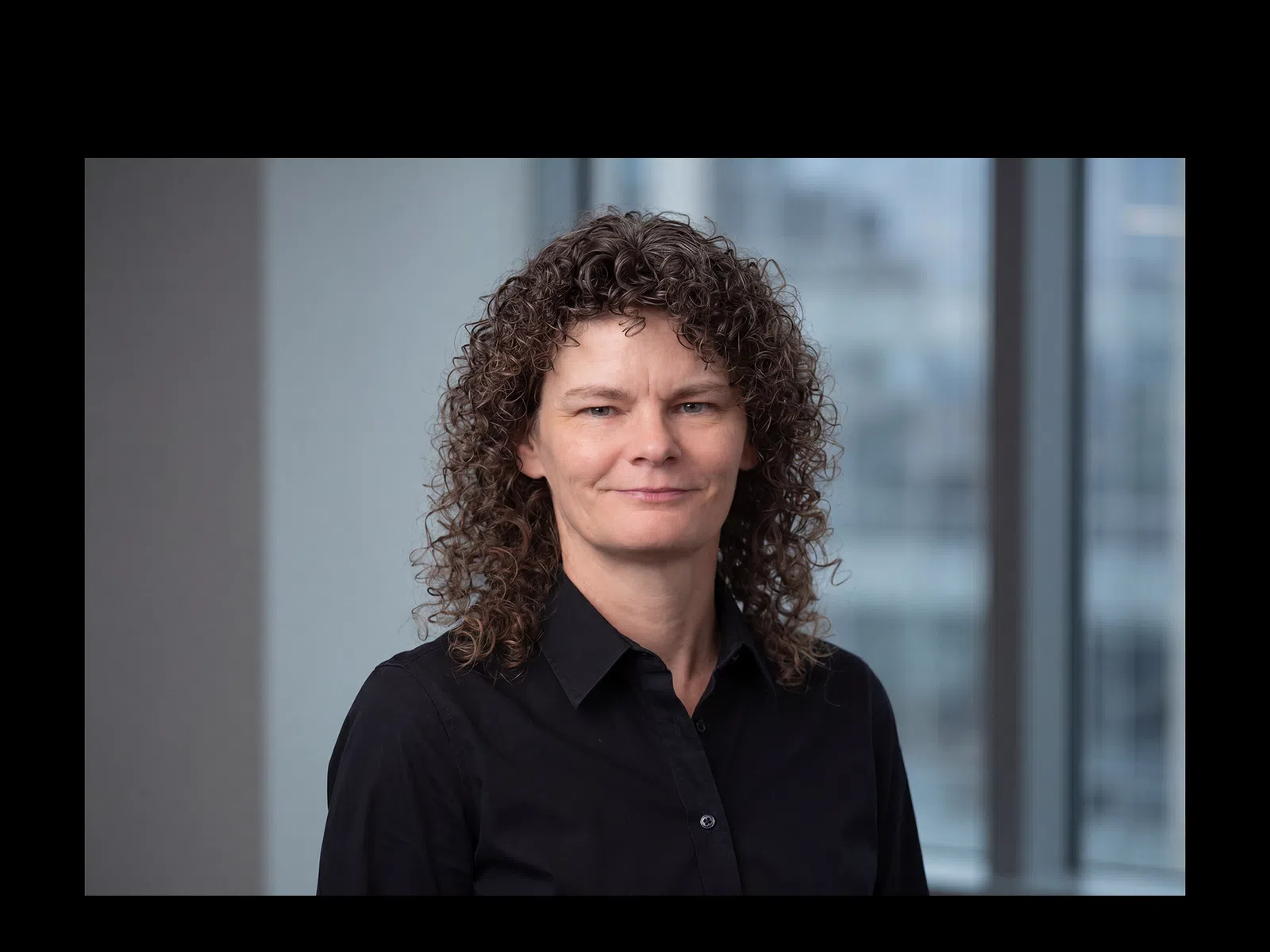 Professional woman with curly hair wearing a black shirt in an office setting with windows behind her