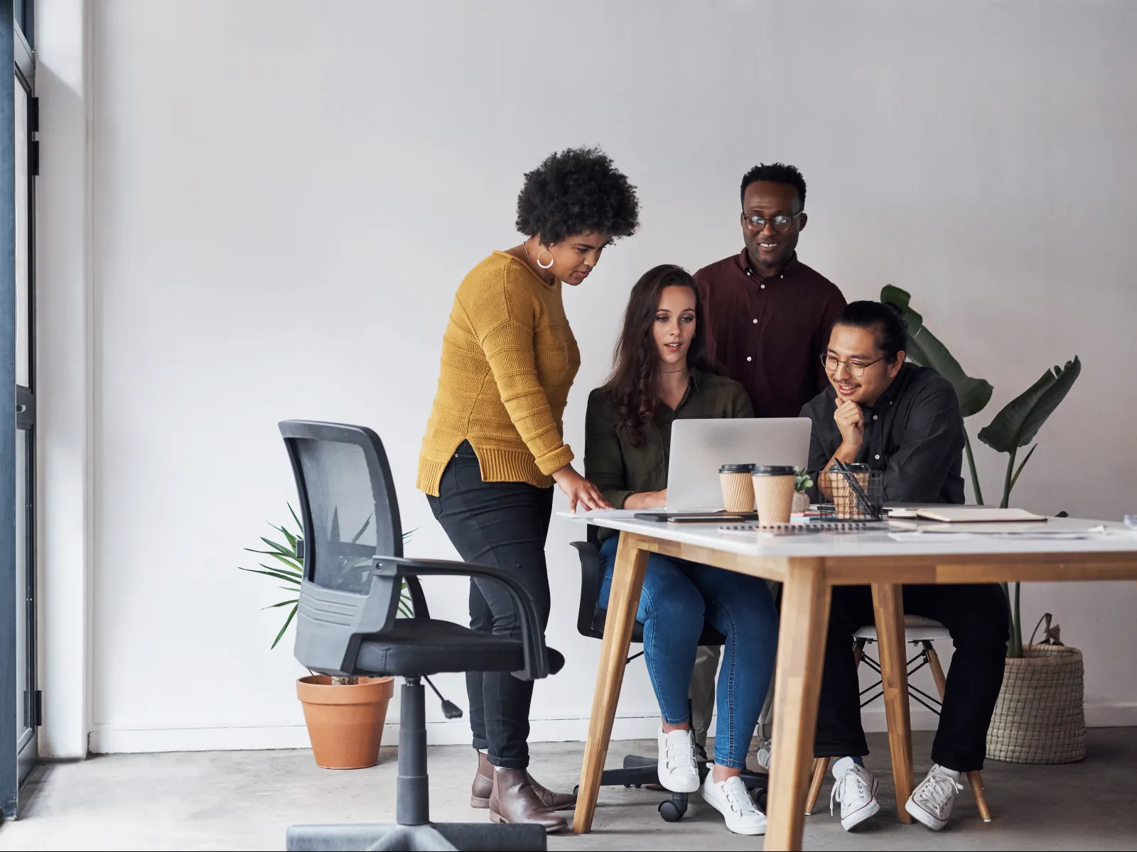 Four diverse colleagues collaborating and discussing work around a laptop in a modern office space.