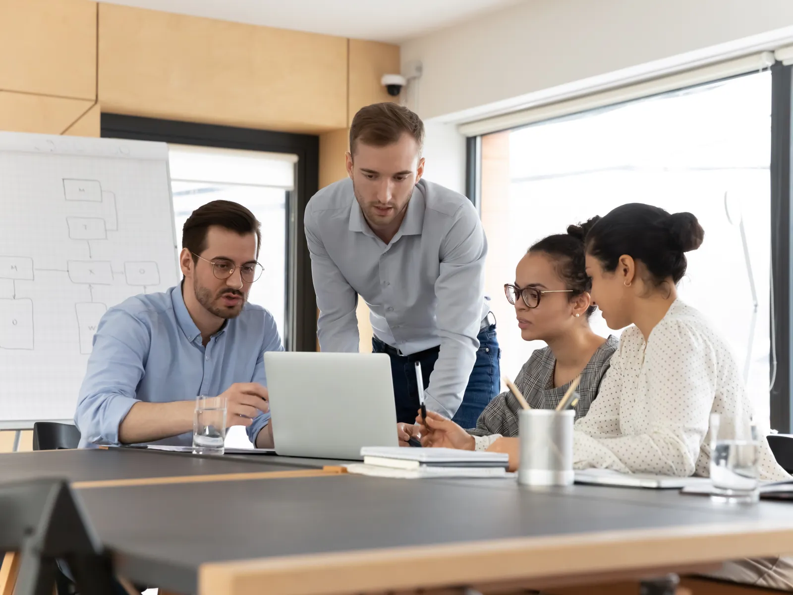 Four diverse colleagues collaborating around a laptop in a modern office meeting room with charts and notebooks.