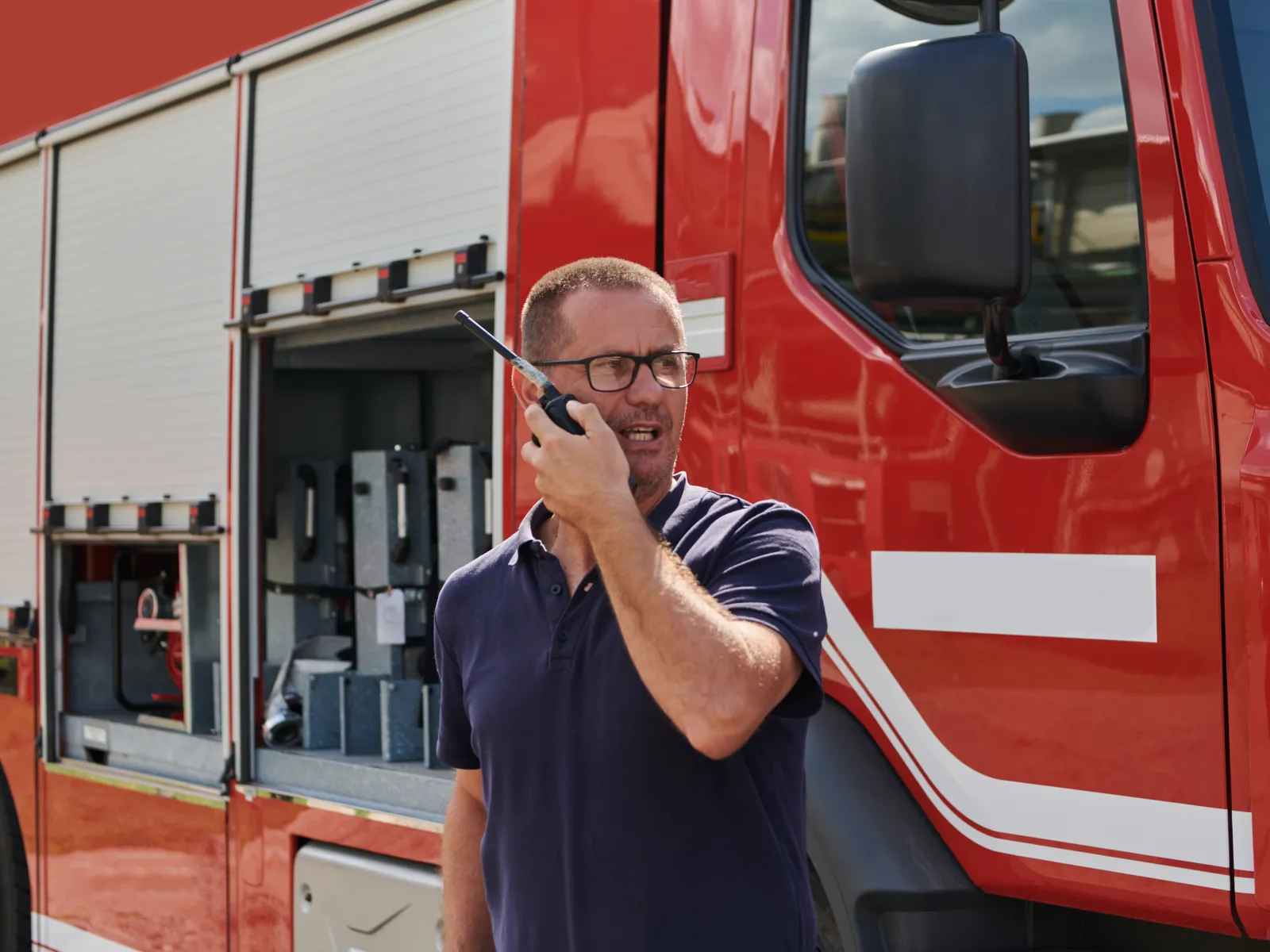 Firefighter in navy shirt using walkie-talkie standing next to red fire truck with open equipment compartments