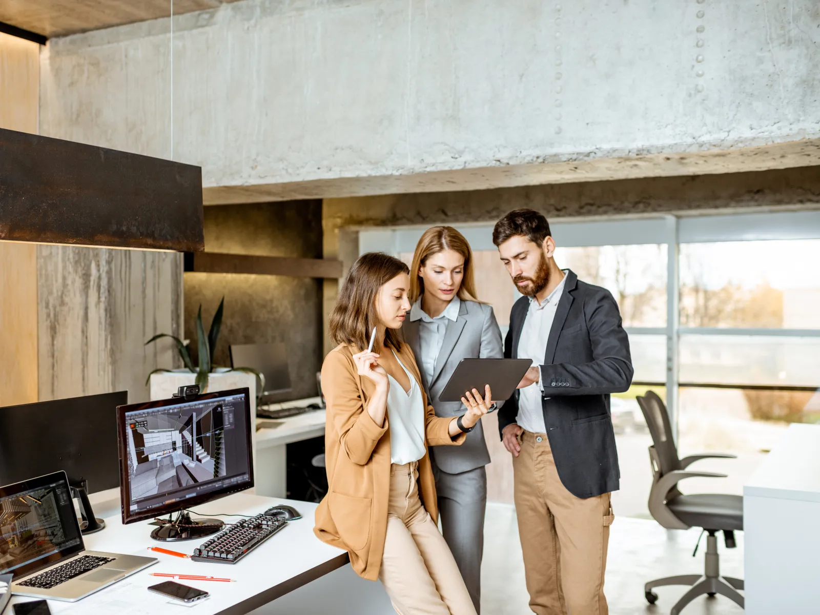 Three professionals collaborating over a tablet in a modern office with computers and architectural designs